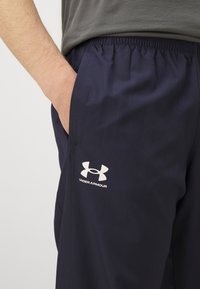Pantalons de sport marins en tissu léger avec une ceinture élastique, présentant un logo blanc Under Armour sur la cuisse gauche.