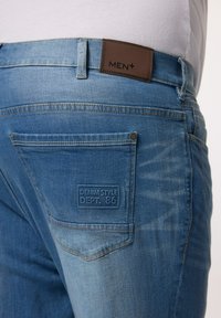 Blå denimjeans med en brun läderetikett märkt "MEN+" på midjan, med en bakficka prägad med "DENIM STYLE DEPT. 86."
