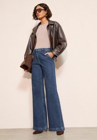 Hnedá kožená bunda, béžový top, široké modré džínsy a hnedé podpätky. Doplnky zahŕňajú veľké slnečné okuliare a hnedú kabelku.