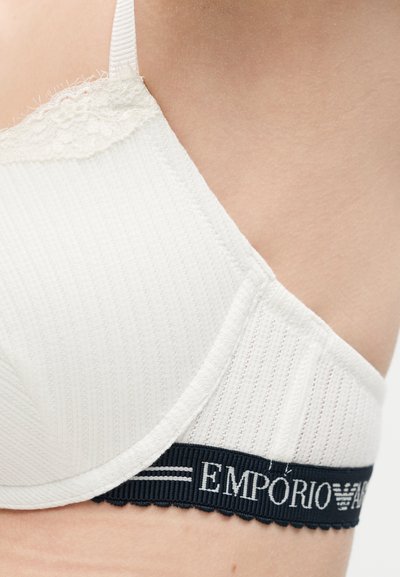 Nærbillede af en person iført en hvid ribstrikket bh med blondestrimler og en sort elastik med teksten "EMPORIO ARMANI."