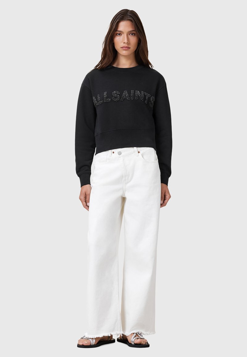Felpa nera cropped con lettering testurizzato "ALL SAINTS", abbinata a jeans bianchi a gamba larga e sandali piatti argentati. Design semplice, linee pulite.