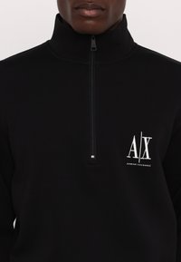 Pullover in pile nero con zip e colletto alto, caratterizzato da un logo bianco ricamato sul lato sinistro del petto e una zip metallica. Texture morbida.