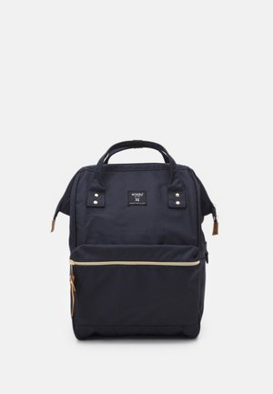 Sac à dos noir de forme carrée avec poignées supérieures, poche zippée frontale et une petite étiquette de marque centrée près du haut.