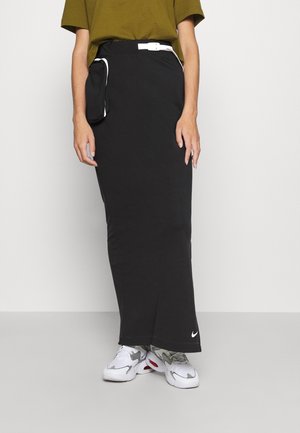 Person iført en lang sort nederdel med sidelomme og hvide sneakers, kombineret med en olivengrøn kortærmet top.
