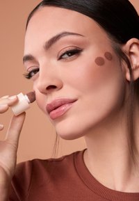 Producto cosmético con aplicador redondeado, aplicando una crema marrón en la mejilla, con una textura suave sobre un fondo neutro.