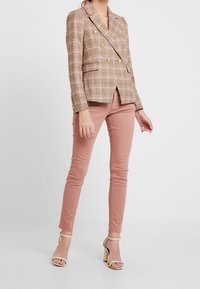 Blazer croisé à motif écossais clair avec des boutons dorés. Associé à un pantalon ajusté rose pastel et des sandales à talons nude.