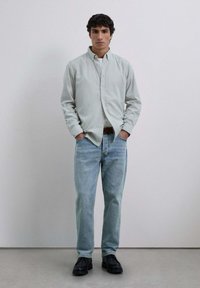 Camisa de manga larga verde claro, con cuello tipo botón, combinada con jeans azul desgastado y zapatos negros de cordones. Diseño simple y casual.