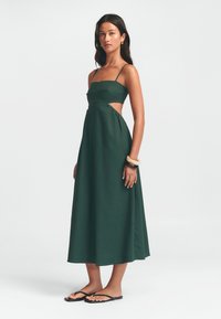 Groene maxi-jurk met spaghettibandjes, uitsparingen bij de taille en een vloeiende rok. Draag met zwarte slippers en een armband.