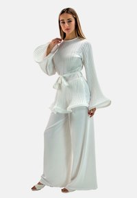 Weißes plissiertes Tunika mit ausgestellt Ärmeln, tailliert mit einem Gürtel, kombiniert mit weit geschnittenen weißen Hosen und minimalistischen Sandalen.