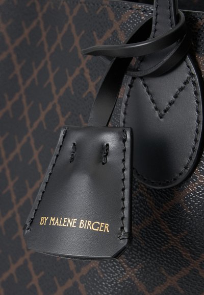 Gros plan sur une étiquette en cuir noir avec un texte doré "BY MALENE BIRGER" sur un fond en cuir marron à motif.