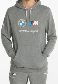 Grijze hoodie van zacht materiaal met een voorkantzak. Heeft BMW en Puma logo's, de tekst "BMW Motorsport" en witte trekkoorden.