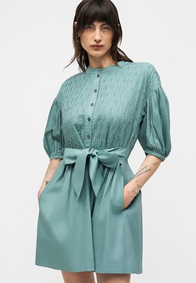 Γυναίκα με σκούρα μαλλιά που φοράει ένα τιρκουάζ, κουμπωτό φόρεμα με υφή στο μπούστο, φουσκωτά μανίκια, μεγάλο φιόγκο στη μέση, με τα χέρια στις τσέπες, στέκεται μπροστά σε λευκό φόντο.