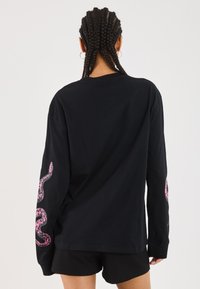 T-shirt noir à manches longues avec un design de serpent rose sur la manche gauche. Fabriqué en coton ; il présente un col rond et une coupe décontractée.