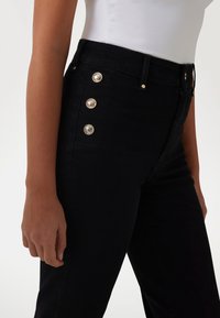 Los jeans de mezclilla negros tienen una cintura alta, cinco botones dorados en el lateral y una forma ajustada, con una textura suave y un diseño minimalista.