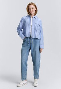 Kratka plava-bijela prugasta košulja na kopčanje s jednim džepom, uparena s širokim svijetloplavim trapericama i bijelim tenisicama.