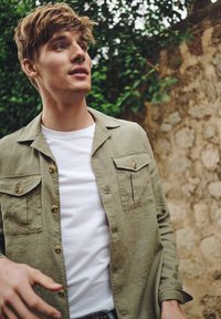 Licht olijf groen overhemdd met twee borstzakken en een wit t-shirt eronder, tegen een achtergrond van een natuurlijke stenen muur.