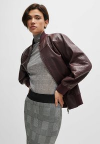 Veste bomber en simili cuir bourgogne avec des poignets et un col côtelés, dotée d'une finition texturée. Associée à un pull à col roulé gris et à une jupe à motifs.