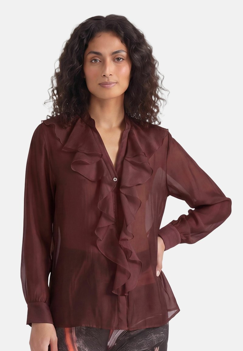 Femme aux cheveux bouclés portant un chemisier bordeaux transparent à manches longues avec des volants et un pantalon à motifs, debout, une main sur la hanche.