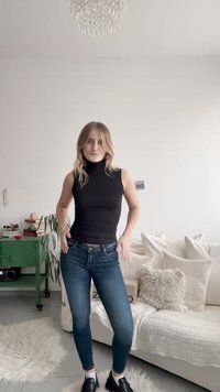 Fekete ujjatlan garbó, sötétkék, szűk szabású farmerrel és fekete belebújós cipővel párosítva. Világos háttér, mellette texturált fehér párnák.