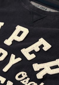 Marineblå sweatshirt med opstående, cremefarvede broderede bogstaver. Rund hals, blød tekstur og et afslappet design. Etiketten synlig indeni.