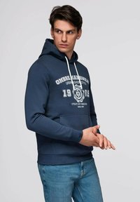 Sweat à capuche bleu marine en tissu doux, avec une poche avant, des cordons blancs et des détails graphiques avec l'année 1985 imprimée.