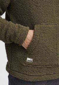 Olive green fleece pullover, textured fabric with a prominent pocket, logo patch on the hem, and elasticated cuffs.