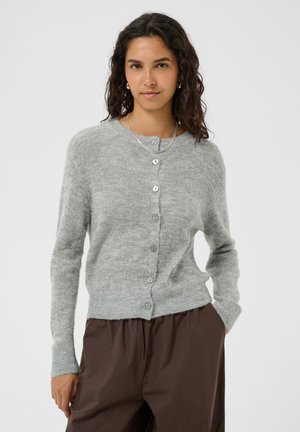 Femme aux cheveux foncés et ondulés, portant un cardigan gris clair boutonné, un pantalon marron, un collier et des boucles d'oreilles en perles, debout devant un fond uni.