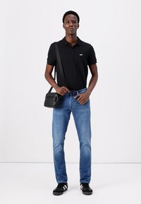 Crna polo majica, plave traperice, crna torba preko tijela i crne tenisice. Model stoji na jednoličnoj pozadini u neutralnom stavu.