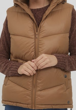 Hnědá péřová vesta na zip s kapucí. Obsahuje prošívané části a druky na spodním lemu. Nosená přes hnědý strukturovaný svetr.