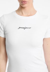 T-shirt blanc à côtes avec un logo "guessjeans" brodé en noir sur la poitrine. Manches courtes et coupe ajustée. Texture lisse.