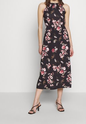 Femme portant une robe midi noire sans manches avec un imprimé floral rose et des sandales noires à talons hauts et brides, debout devant un fond uni.