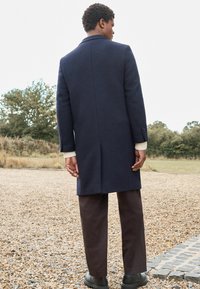 Abrigo de lana azul marino con una silueta larga, ventilación trasera simple y puños con botones, combinado con pantalones rectos marrones y zapatos negros.