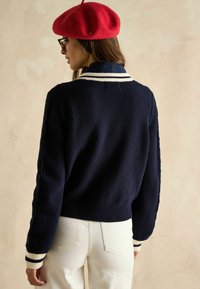 Jūras spēku teksturēts džemperis ar baltu svītru apkakli un piedurknēm, ar kabeli adītām piedurknēm. Modelis valkā baltas bikses un sarkanu bereti.