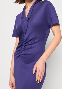 Fialové šaty s krátkým rukávem mají obepínající výstřih, nařasení v pase, hladkou texturu a přiléhavý střih.
