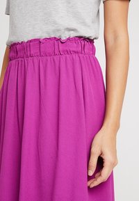 Rosa, flödande kjol i strukturerad tyg med en samlad, elastisk midja, visad med en hand som vilar på tyget.