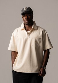 Polo shirt beige con zip, colletto, maniche corte e taschino con un accento dorato. Cappellino da baseball grigio con logo.