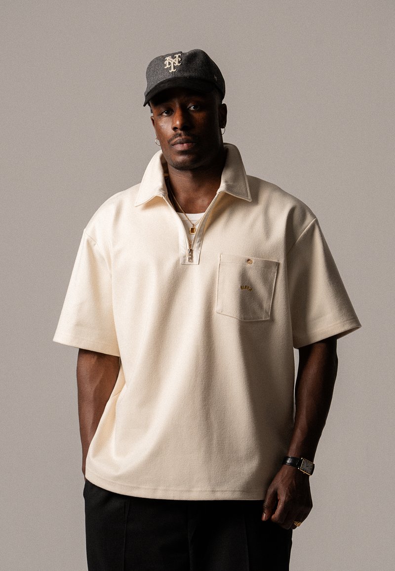 Polo shirt beige con zip, colletto, maniche corte e taschino con un accento dorato. Cappellino da baseball grigio con logo.