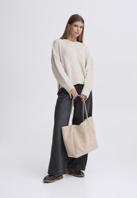 Pulover gros din tricot crem cu model diagonal în coaste, asortat cu blugi evazați închisi la culoare și o geantă tote moale bej. Încălțămintea are tălpi groase și maro.