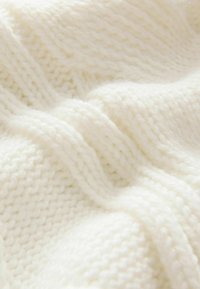 Wollen deken in crème, met een gevarieerd gebreid patroon van verschillende steken, zacht en dik materiaal, zachtjes gedrapeerd.