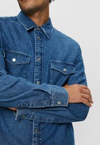 Camisa de denim en azul medio con un cuello clásico, dos bolsillos en el pecho y puños con botón. Presenta botones de metal y un corte relajado.