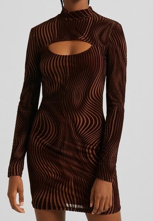 Femme portant une mini-robe marron à manches longues avec un motif rayé ondulé et une découpe sous l'encolure sur un fond uni.