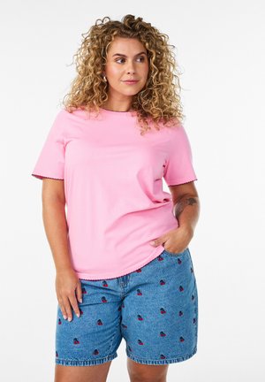 Nainen, jolla on kiharat hiukset, pukeutunut vaaleanpunaiseen lyhythihaiseen paitaan ja sinisiin farkkushortseihin, joissa on pieniä kirsikkakuvioita, toinen käsi taskussa, neutraali tausta.