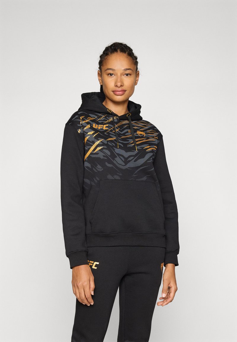 Sweatshirt preto com uma sobreposição texturizada em padrões dourados e cinzentos, bolso canguru e capuz com cordão; combinado com calças de moletom pretas a condizer.