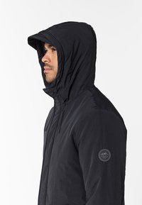 Chaqueta negra con capucha de textura suave, que presenta un parche con el logo en la manga izquierda y cordones en la capucha para ajustarla.
