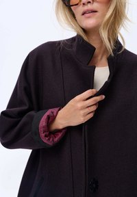 Donkerpaars oversized jas met een hoge kraag, contrasterende textuur van zwarte manchetten en een bordeauxrode met patronen gevoerde binnenkant, met een knoopsluiting aan de voorkant.