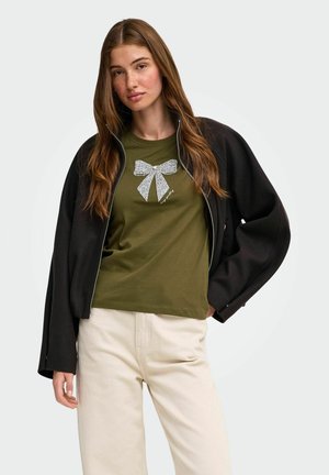 Ung kvinde med langt brunt hår iført en sort åben jakke, grøn t-shirt med et sølv sløjfe-design og lyse beige bukser.
