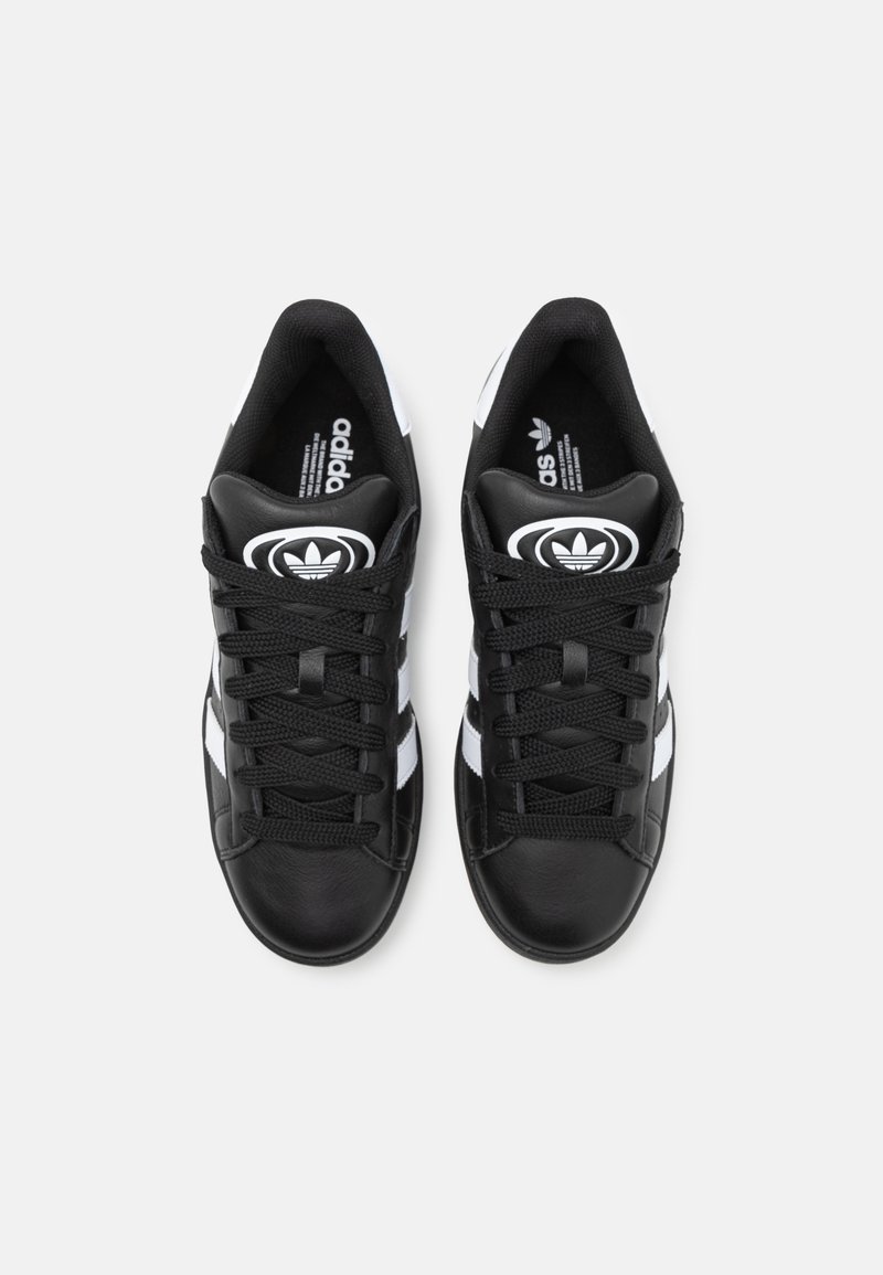Zapatillas de cuero negro con rayas blancas, logo redondo negro y cordones negros. El diseño es bajo con una textura suave.