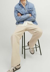 Světle modrá džínová bunda se dvěma náprsními kapsami, oblečená přes bílou košili. Béžové kalhoty a šedé tenisky s černými akcenty doplňují outfit.