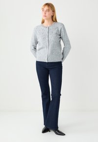 Grijze wollen cardigan met een ronde hals, versierd met decoratieve kralen aan de voorkant, gecombineerd met uitlopende donkere spijkerbroek en zwarte puntige schoenen.