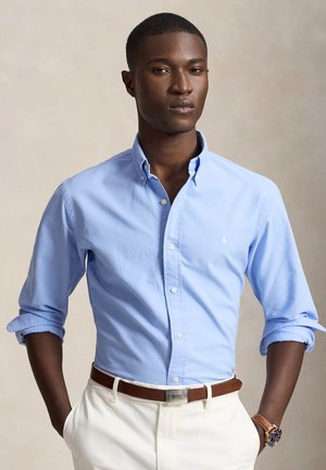 Jeune homme portant une chemise bleu clair à boutons avec les manches retroussées, un pantalon blanc, une ceinture marron et une montre au poignet, debout les mains dans les poches.